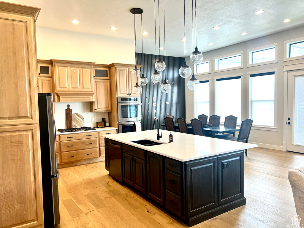 Kitchen featuring appliances with stainless steel finishes, light hardwood / wood-style floors, sink, pendant lighting, and an island with sink