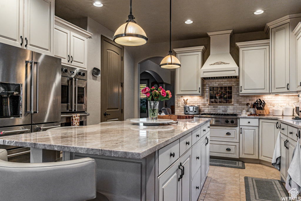 Kitchen featuring stainless steel appliances, a kitchen island, custom exhaust hood, light tile flooring, and backsplash