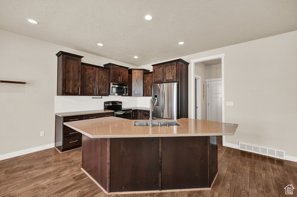 Kitchen featuring dark wood-type flooring, appliances with stainless steel finishes, dark brown cabinets, a kitchen island with sink, and light stone countertops