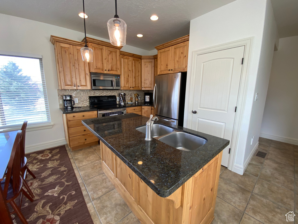 Kitchen with decorative light fixtures, appliances with stainless steel finishes, an island with sink, light tile floors, and tasteful backsplash