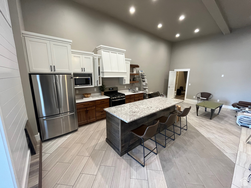 Kitchen featuring appliances with stainless steel finishes, a towering ceiling, white cabinets, light stone countertops, and a kitchen island with sink