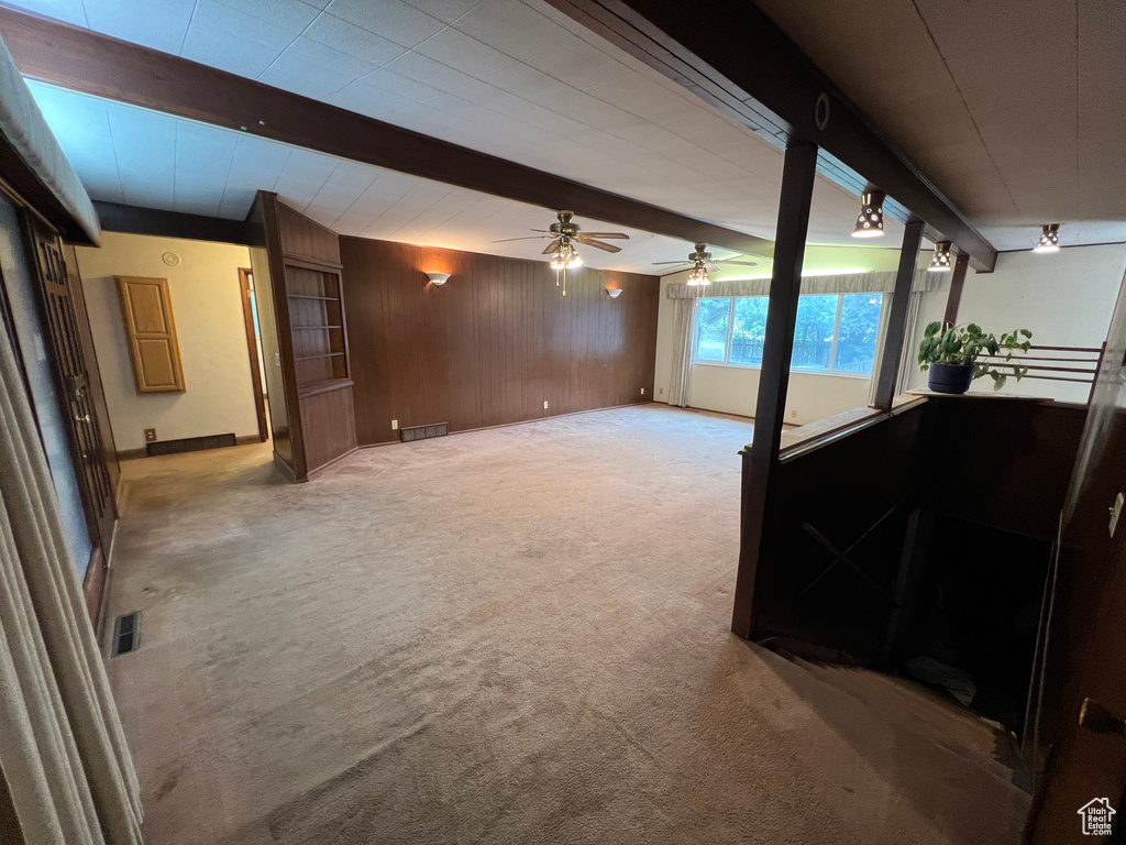 Unfurnished living room featuring light colored carpet, wood walls, ceiling fan, and beam ceiling