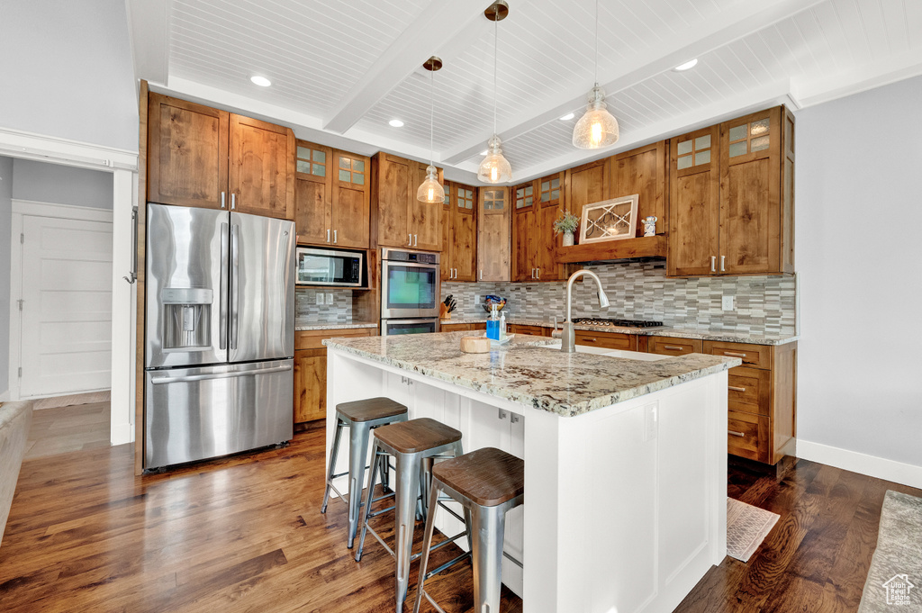 Kitchen with decorative light fixtures, beamed ceiling, appliances with stainless steel finishes, a center island with sink, and tasteful backsplash