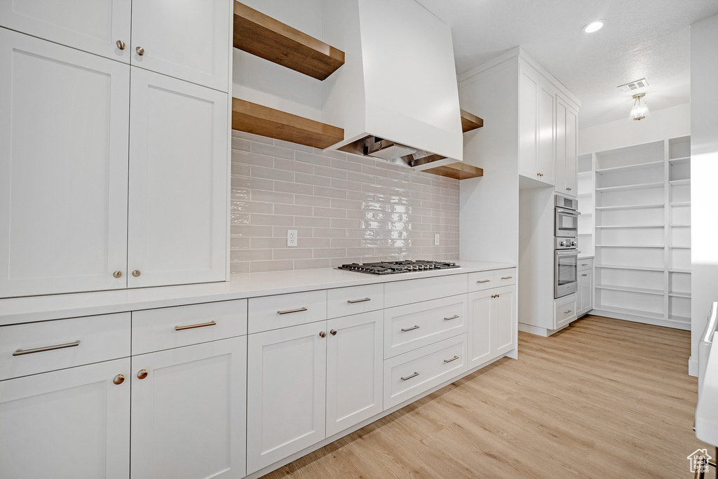 Kitchen featuring custom range hood, light wood-type flooring, tasteful backsplash, stainless steel appliances, and white cabinetry