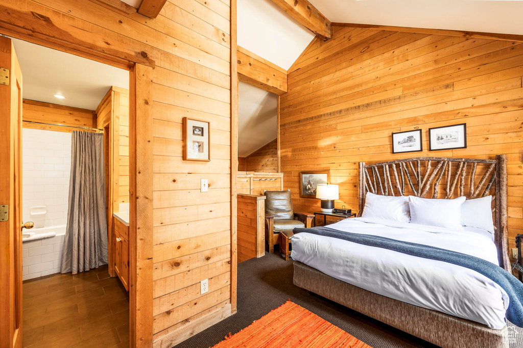 Carpeted bedroom featuring vaulted ceiling with beams, ensuite bathroom, and wooden walls