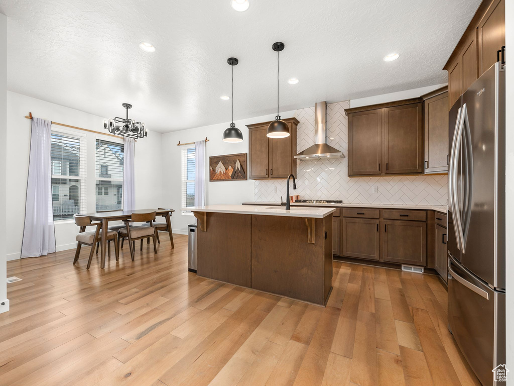 Kitchen featuring hanging light fixtures, light hardwood / wood-style flooring, a center island with sink, stainless steel appliances, and wall chimney exhaust hood