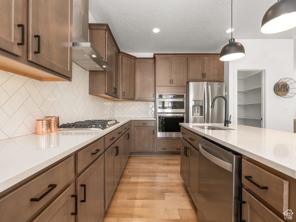 Kitchen with light hardwood / wood-style floors, stainless steel appliances, tasteful backsplash, wall chimney exhaust hood, and hanging light fixtures