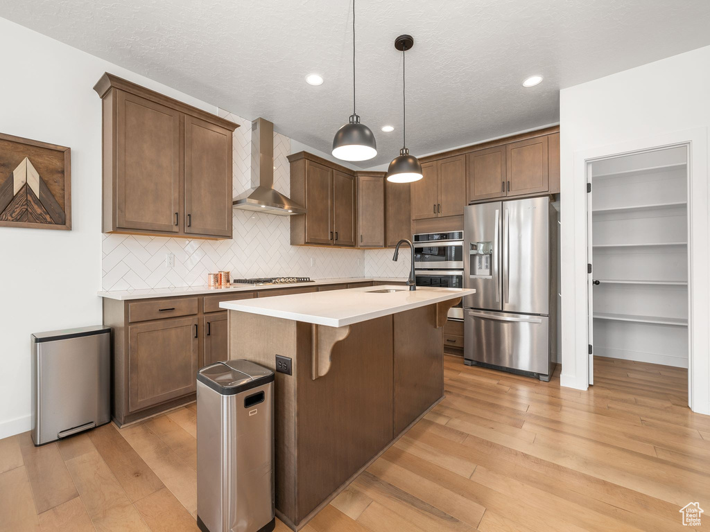 Kitchen featuring a kitchen island with sink, pendant lighting, light hardwood / wood-style floors, stainless steel appliances, and wall chimney exhaust hood