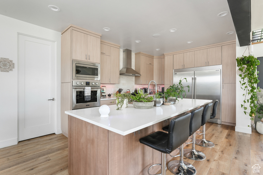 Kitchen featuring light hardwood / wood-style flooring, a breakfast bar area, built in appliances, wall chimney exhaust hood, and a center island with sink