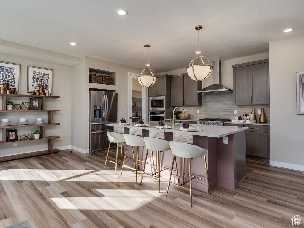 Kitchen featuring appliances with stainless steel finishes, a kitchen island with sink, wall chimney range hood, and light hardwood / wood-style flooring