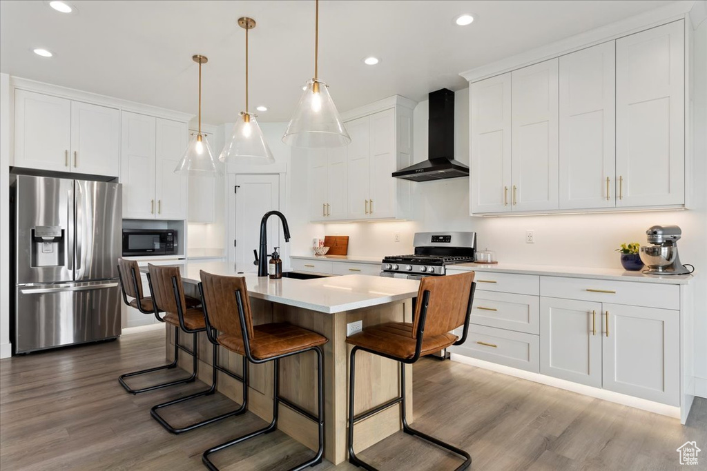 Kitchen with decorative light fixtures, appliances with stainless steel finishes, light hardwood / wood-style floors, white cabinets, and wall chimney exhaust hood