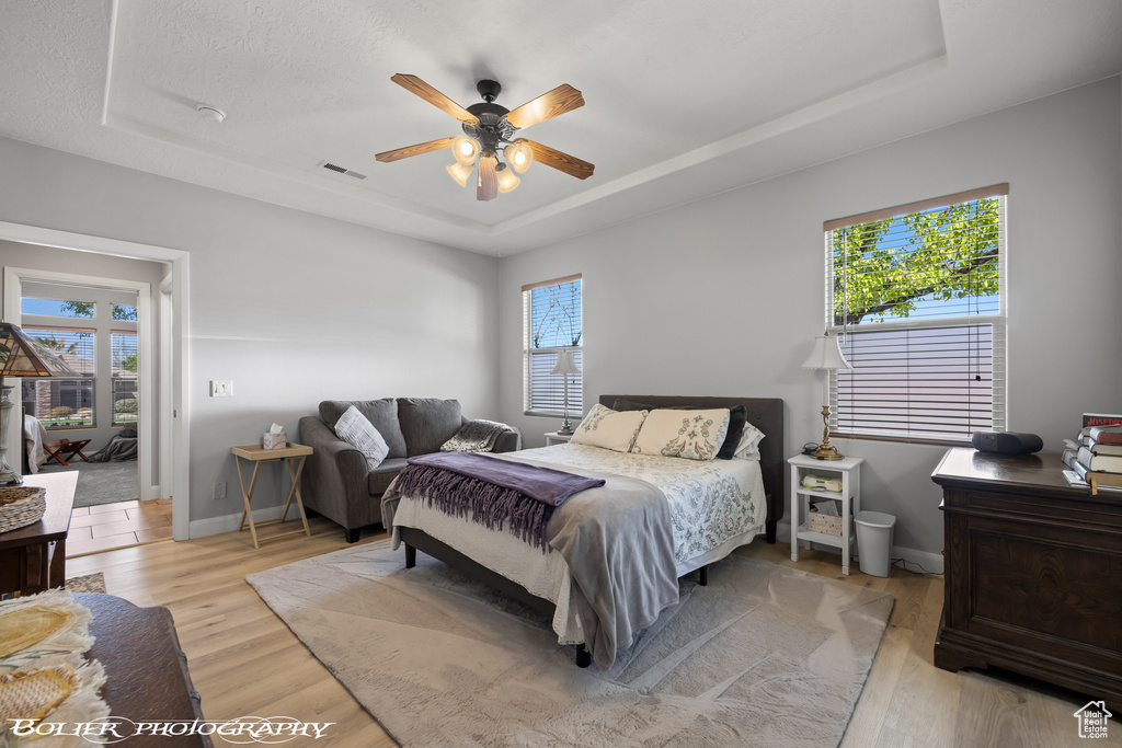 Bedroom featuring multiple windows, light hardwood / wood-style flooring, a tray ceiling, and ceiling fan