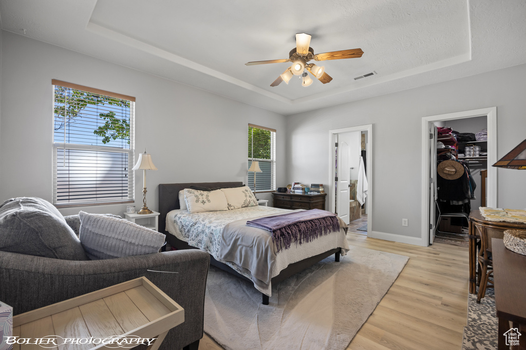 Bedroom with ensuite bathroom, ceiling fan, a spacious closet, light hardwood / wood-style flooring, and a tray ceiling
