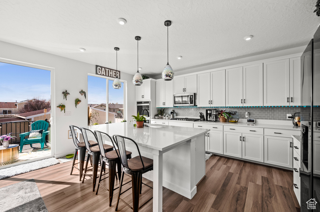 Kitchen featuring stainless steel appliances, decorative light fixtures, a center island with sink, white cabinetry, and dark hardwood / wood-style flooring