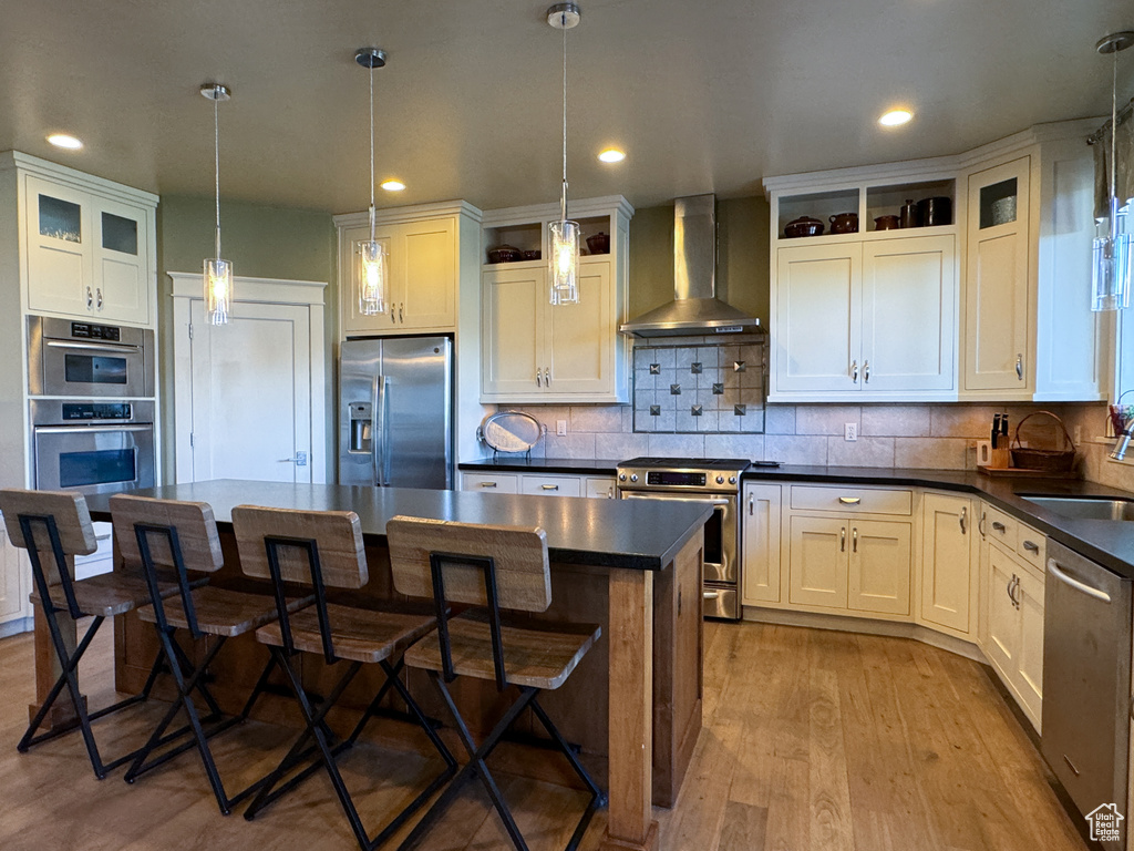 Kitchen featuring appliances with stainless steel finishes, light hardwood / wood-style floors, a kitchen island, pendant lighting, and wall chimney range hood
