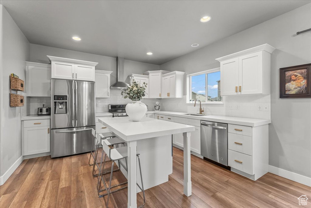 Kitchen featuring appliances with stainless steel finishes, backsplash, light hardwood / wood-style flooring, wall chimney exhaust hood, and white cabinets