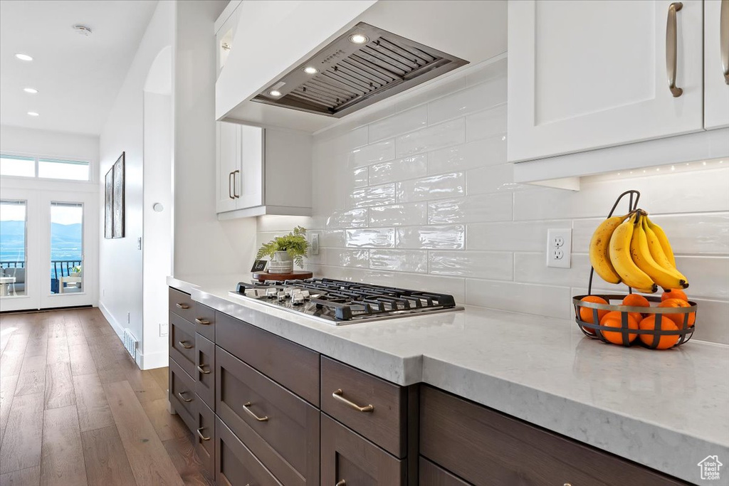 Kitchen featuring dark wood-type flooring, wall chimney range hood, backsplash, dark brown cabinetry, and white cabinets