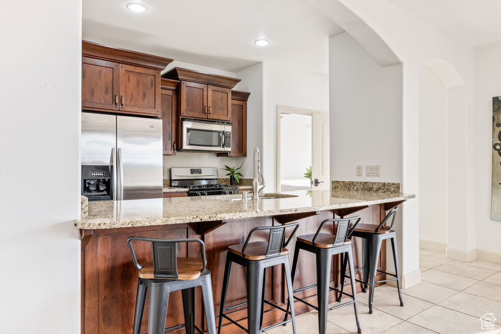 Kitchen featuring appliances with stainless steel finishes, light tile flooring, a kitchen breakfast bar, kitchen peninsula, and light stone countertops