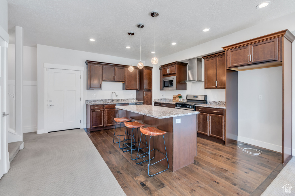 Kitchen featuring dark wood-type flooring, appliances with stainless steel finishes, pendant lighting, wall chimney range hood, and a kitchen island