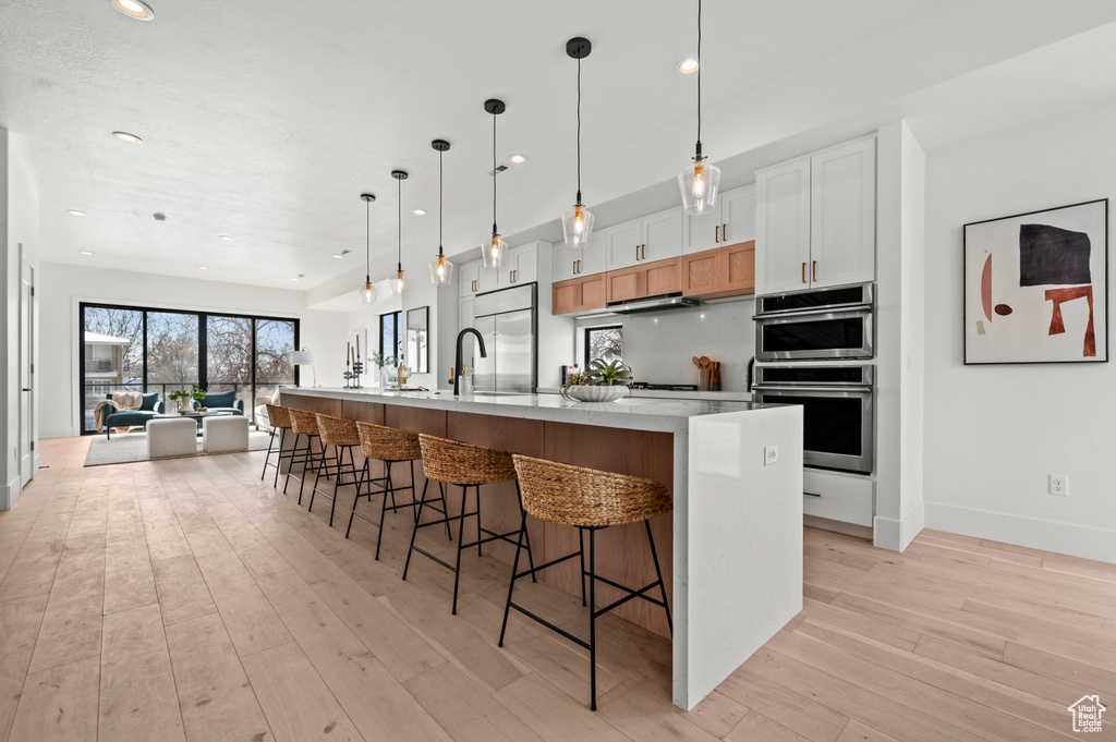 Kitchen featuring stainless steel appliances, white cabinets, light hardwood / wood-style flooring, an island with sink, and decorative light fixtures