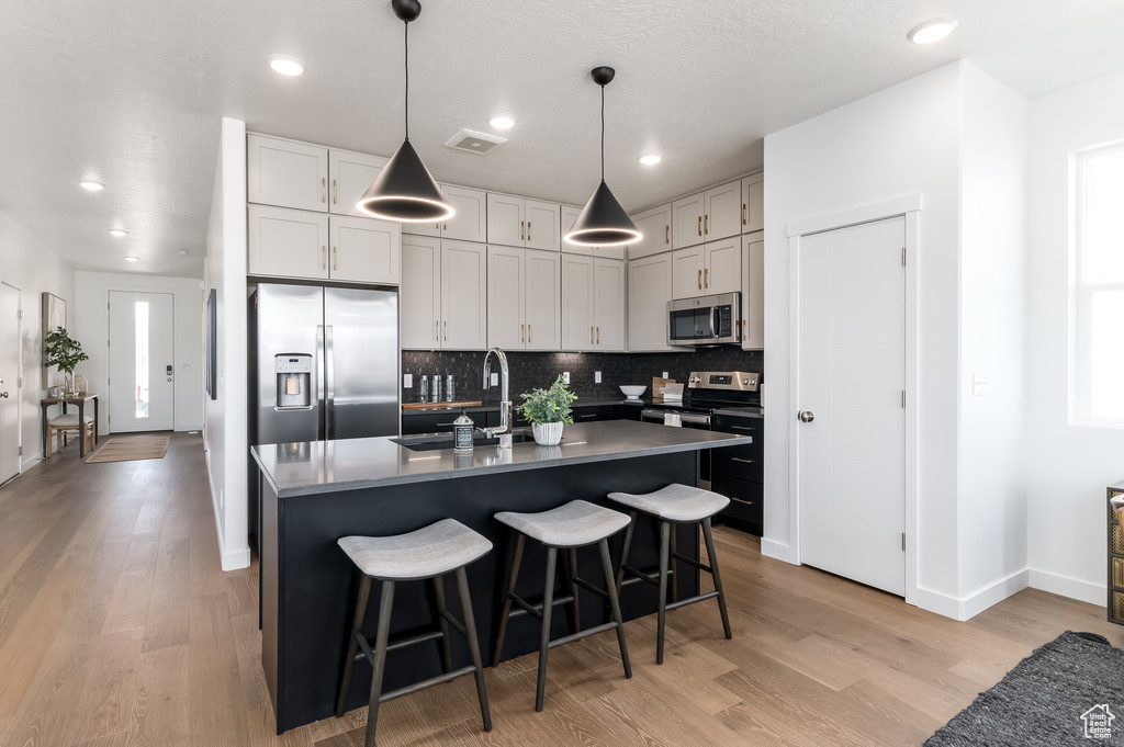 Kitchen featuring stainless steel appliances, backsplash, pendant lighting, light hardwood / wood-style floors, and a center island with sink