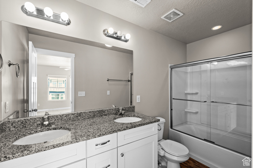 Full bathroom featuring shower / bath combination with glass door, toilet, a textured ceiling, and dual bowl vanity
