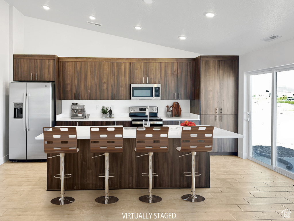 Kitchen featuring appliances with stainless steel finishes, dark brown cabinetry, a kitchen island with sink, a kitchen breakfast bar, and vaulted ceiling