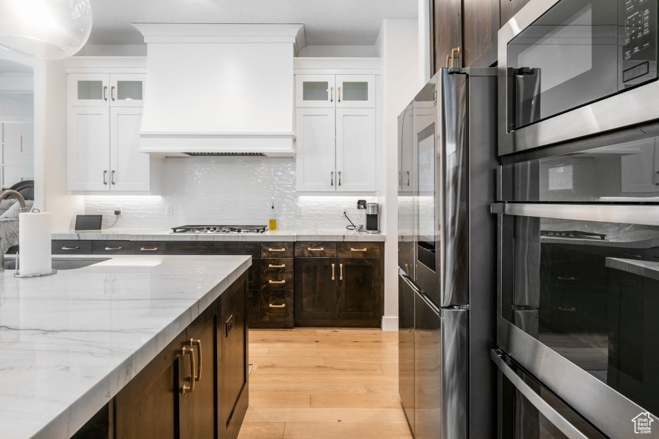 Kitchen featuring backsplash, appliances with stainless steel finishes, premium range hood, white cabinetry, and light wood-type flooring