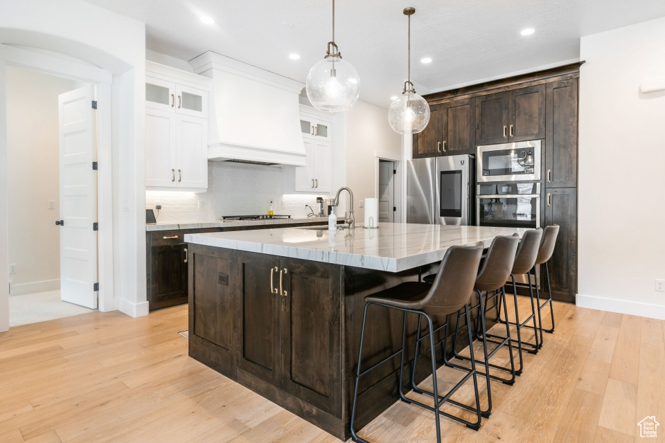 Kitchen featuring appliances with stainless steel finishes, backsplash, custom exhaust hood, light hardwood / wood-style flooring, and a kitchen island with sink