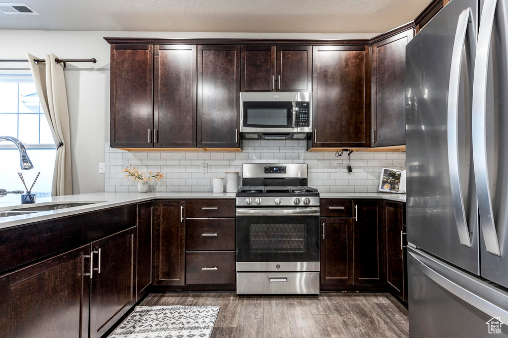 Kitchen featuring appliances with stainless steel finishes, tasteful backsplash, dark brown cabinets, wood-type flooring, and sink