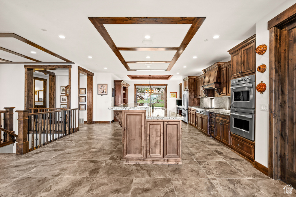 Kitchen with an inviting chandelier, light stone countertops, hanging light fixtures, stainless steel appliances, and a kitchen island with sink