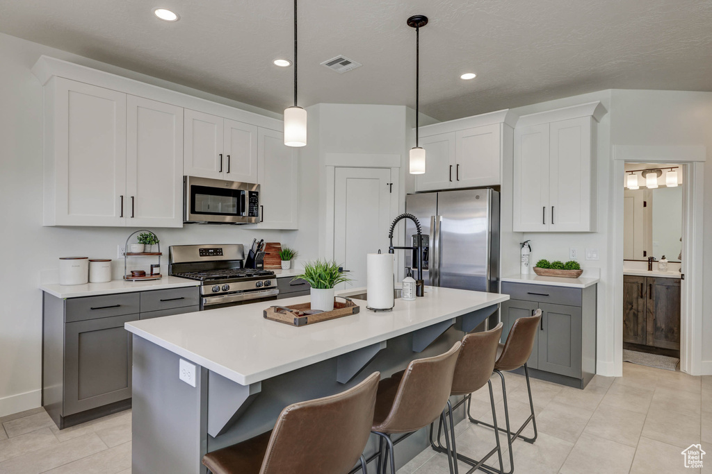 Kitchen with gray cabinets, hanging light fixtures, appliances with stainless steel finishes, a center island with sink, and a kitchen bar