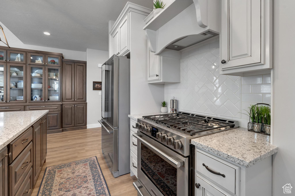 Kitchen with tasteful backsplash, white cabinetry, premium appliances, light hardwood / wood-style flooring, and premium range hood