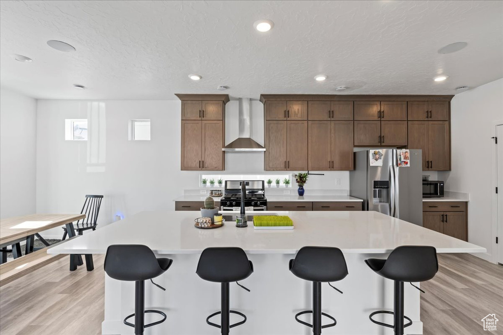 Kitchen with wall chimney exhaust hood, a center island with sink, light hardwood / wood-style floors, and stainless steel refrigerator with ice dispenser