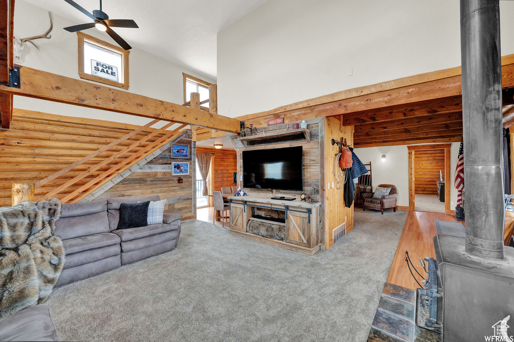 Carpeted living room with a wood stove, wooden walls, ceiling fan, log walls, and a high ceiling