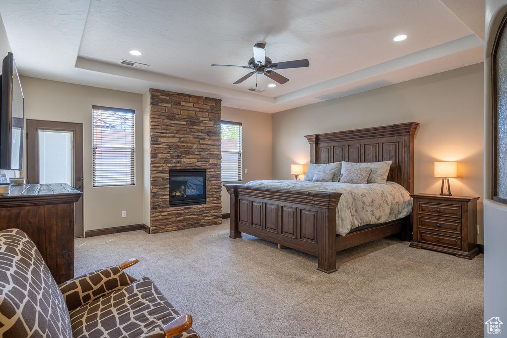 Carpeted bedroom featuring ceiling fan, a tray ceiling, and a stone fireplace