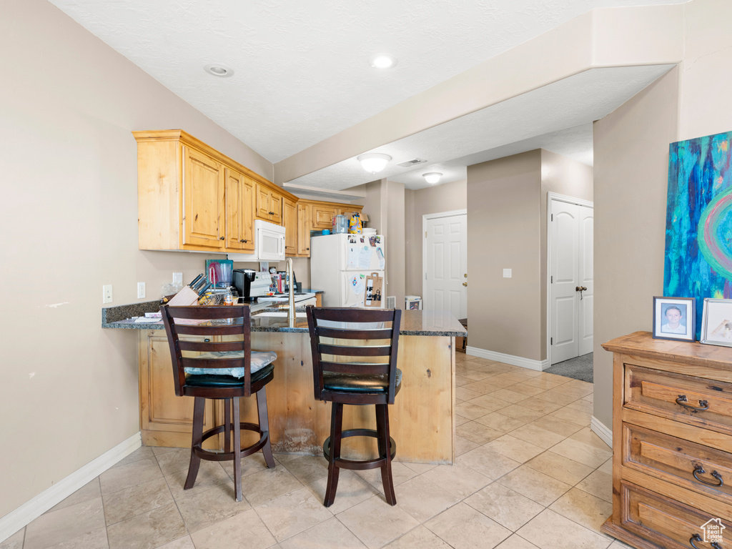 Kitchen featuring a breakfast bar, white appliances, kitchen peninsula, light tile floors, and dark stone countertops