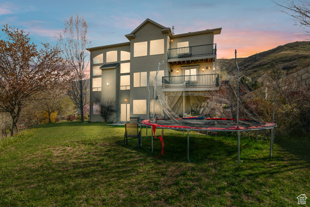 Back house at dusk featuring a trampoline, a balcony, and a lawn