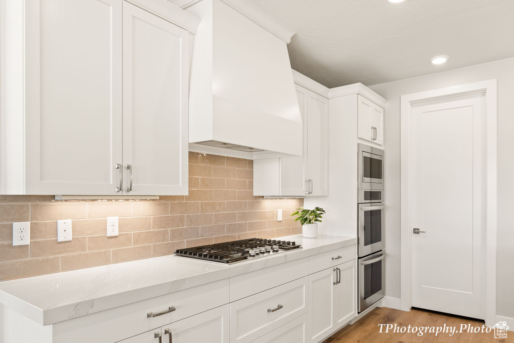 Kitchen featuring custom exhaust hood, white cabinetry, backsplash, appliances with stainless steel finishes, and light hardwood / wood-style floors