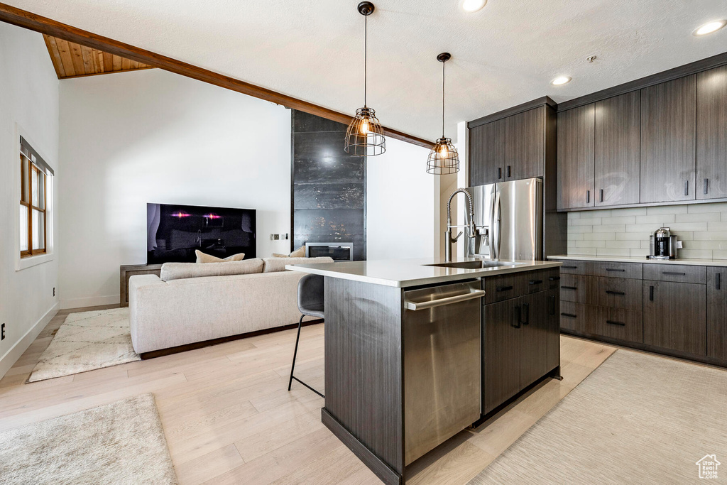 Kitchen with light wood-type flooring, appliances with stainless steel finishes, dark brown cabinets, a center island with sink, and tasteful backsplash
