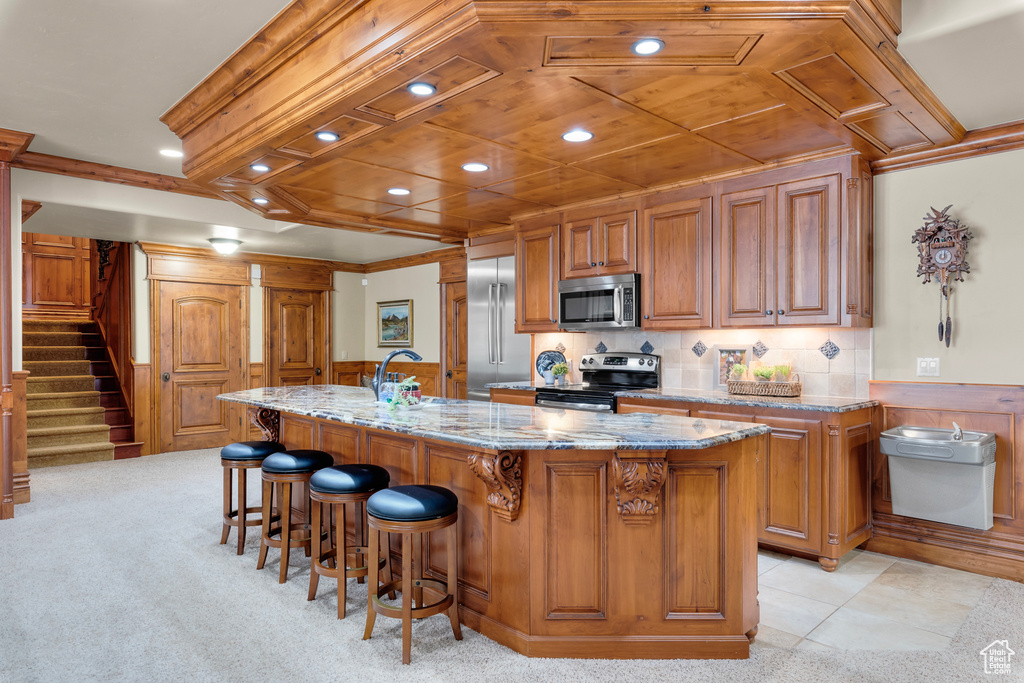 Kitchen with a kitchen island with sink, light stone countertops, stainless steel appliances, light tile floors, and a kitchen bar