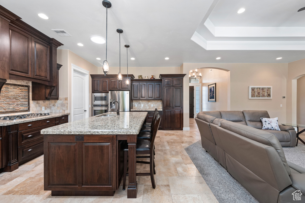 Kitchen featuring stainless steel appliances, tasteful backsplash, an island with sink, light stone counters, and dark brown cabinetry