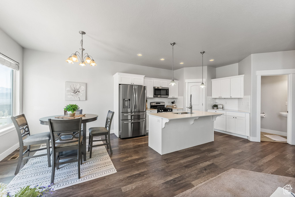 Kitchen with appliances with stainless steel finishes, white cabinetry, dark wood-type flooring, hanging light fixtures, and a kitchen island with sink