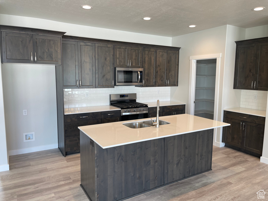 Kitchen featuring backsplash, appliances with stainless steel finishes, light hardwood / wood-style floors, and an island with sink