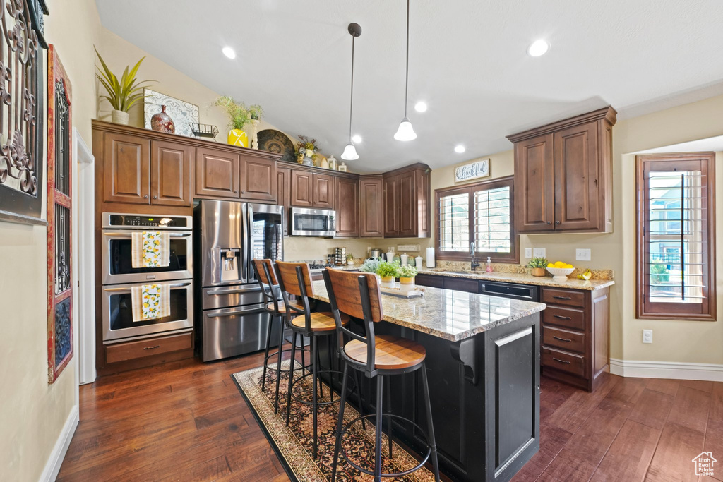 Kitchen with decorative light fixtures, dark wood-type flooring, appliances with stainless steel finishes, light stone counters, and a kitchen island