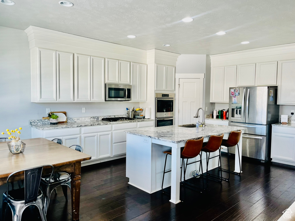 Kitchen featuring light stone countertops, appliances with stainless steel finishes, a kitchen island with sink, white cabinetry, and dark hardwood / wood-style flooring