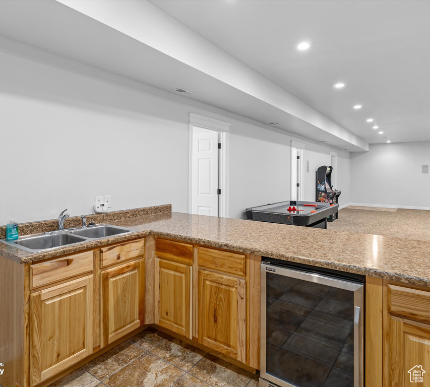 Kitchen featuring kitchen peninsula, light tile flooring, wine cooler, light stone counters, and sink