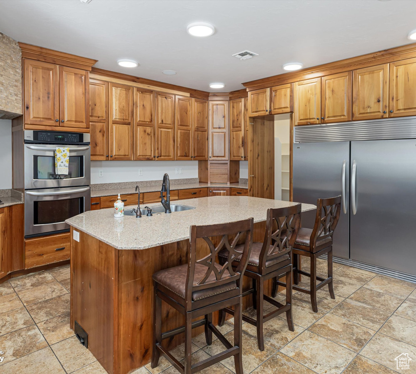 Kitchen featuring a center island with sink, appliances with stainless steel finishes, a kitchen breakfast bar, and light stone counters