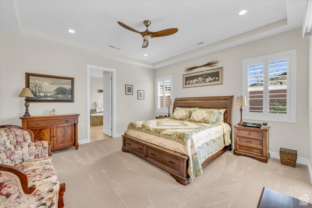 Carpeted bedroom with connected bathroom, ceiling fan, a raised ceiling, and multiple windows