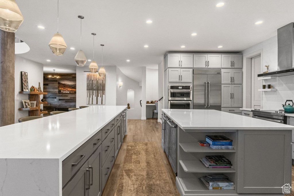 Kitchen with a kitchen island, gray cabinets, stainless steel appliances, wall chimney range hood, and pendant lighting